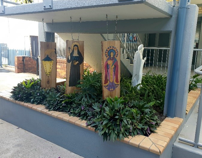 Outdoor garden with painted saint panels and a white statue set among plants near school buildings