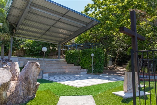 Covered outdoor prayer space with a wooden cross, seating area and surrounding greenery