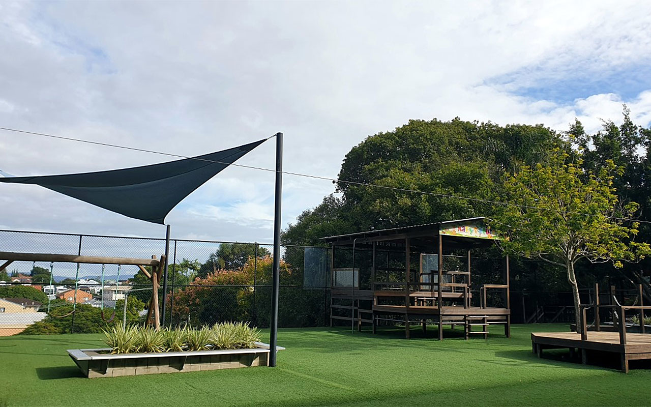 Playground area with shade sail, garden beds and timber seating on grass