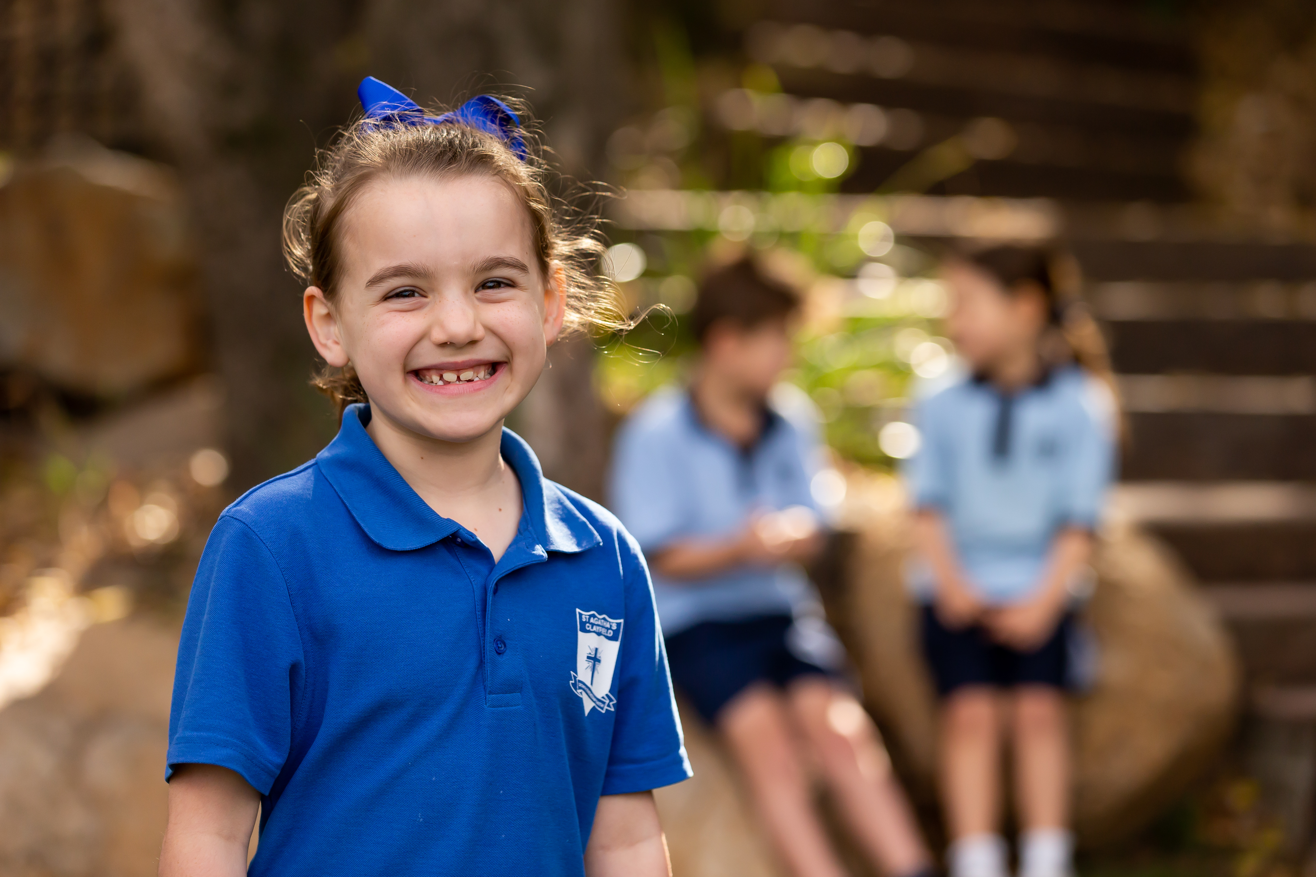 Student in blue school uniform smiling outdoors with classmates blurred in the background