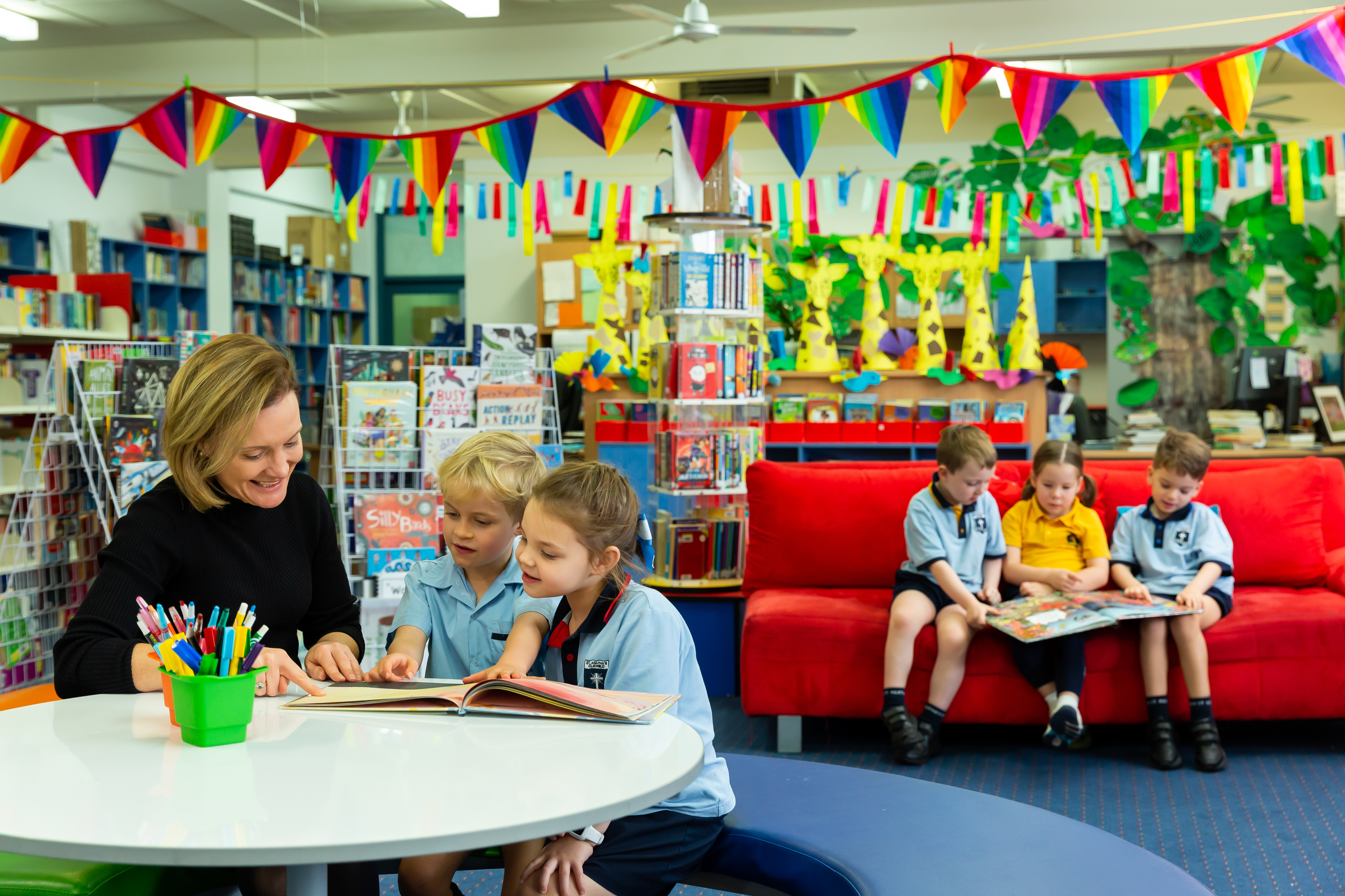 Teacher reading a book with students at a table in the school library, with bookshelves, colourful bunting and other students reading in the background