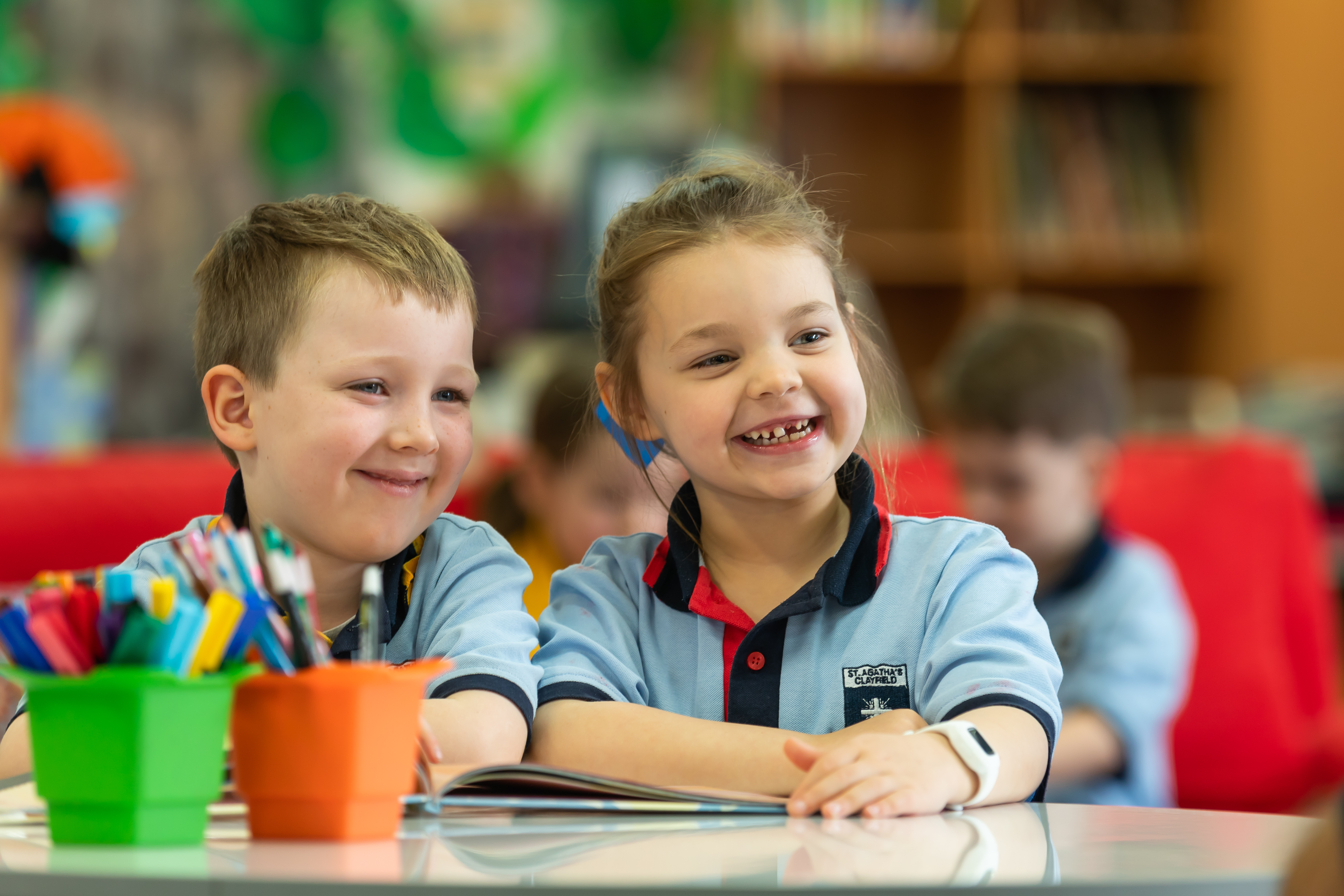 Two young students in school uniforms smiling while sitting together at a classroom table with pencils and books