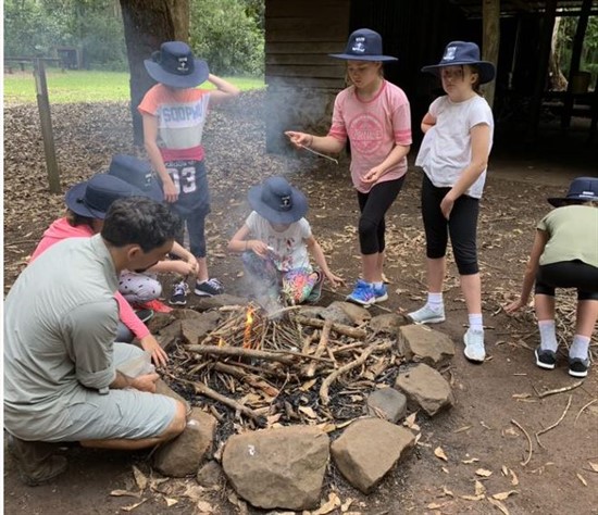 Students gathered around a campfire with a teacher, learning outdoor survival and teamwork skills