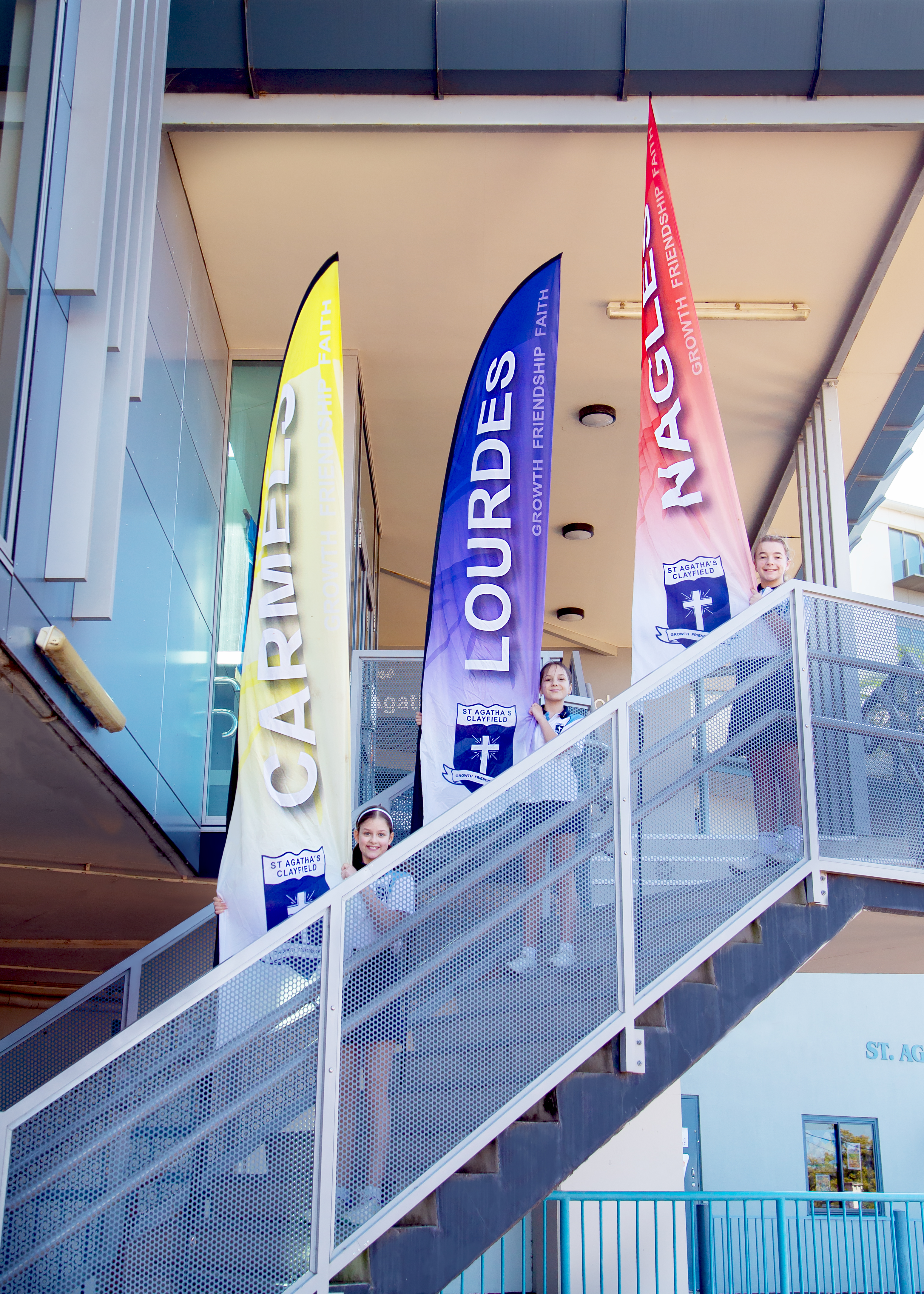 Students standing on an external stairway holding vertical school banners displaying caritas, lourdes and nano nagle with school values text